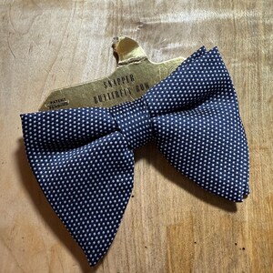 May include: A navy blue bow tie with white polka dots. The bow tie is on a gold card with the text "Snapper Butterfly Bow" and "Patent Pending".
