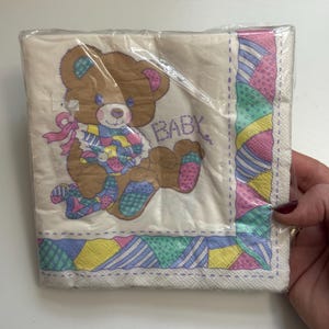 May include: A package of baby-themed napkins. The napkins feature a brown teddy bear with colorful patchwork clothing and the word "BABY". The border has a patchwork design in pastel colors.