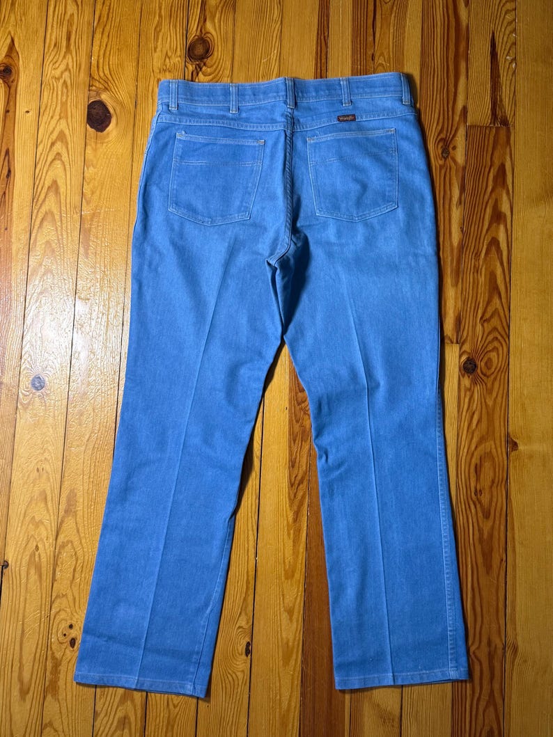May include: A pair of blue denim jeans with a bootcut leg. The jeans have a brown leather patch on the back with the word "Wrangler" printed on it.