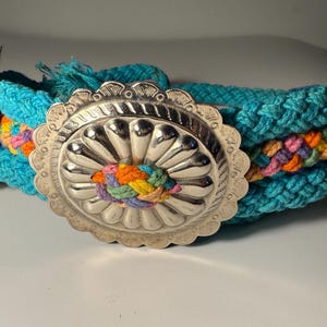 Op de afbeelding: Een turquoise blauwe gevlochten touwriem met een zilveren concho gesp. De concho heeft een reliëfdesign en is omgeven door een decoratieve rand. Het touw is gevlochten met kleurrijke draden.