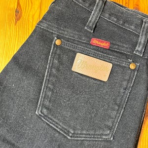 May include: Black denim jeans with a leather Wrangler patch on the back pocket. The patch has the word "Wrangler" in gold lettering.
