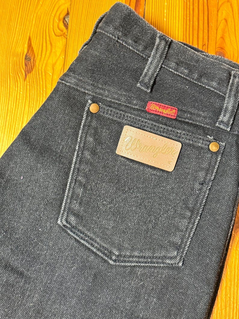 May include: Black denim jeans with a leather Wrangler patch on the back pocket. The patch has the word "Wrangler" in gold lettering.