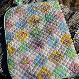 Könnte beinhalten: Ein gesteppter Wandbehang aus Stoff mit einem sich wiederholenden Rautenmuster in den Pastellfarben Gelb, Blau, Rosa und Grün. Jedes Quadrat enthält eine gedruckte Illustration einer Figur mit Wollhaaren und Luftballons.