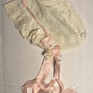 Könnte beinhalten: Elfenbeinfarbene Babyhaube mit Lochmuster und gewelltem Rand. Die Haube hat ein rosa Satinband zum Binden unter dem Kinn. Ein Knopfverschluss ist an der Seite sichtbar. Diese Haube im Vintage-Stil ist für ein Baby.