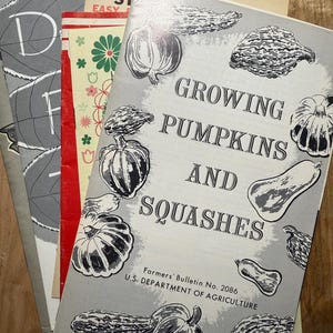 May include: A stack of vintage gardening books with a gray cover featuring black and white illustrations of pumpkins and squashes. The title of the book is "Growing Pumpkins and Squashes" and the text "Farmers' Bulletin No. 2086 U.S. DEPARTMENT OF AGRICULTURE" is printed on the bottom of the cover.