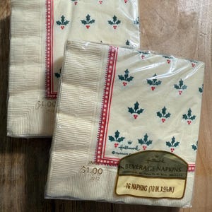 May include: Two sealed packages of Hallmark beverage napkins. The napkins are cream-colored with a pattern of green holly leaves and red berries. Each package has a red border with white dots and the text "16 NAPKINS (10 IN. X 9 5/8 IN.)".