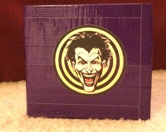 Könnte beinhalten: Eine lila Box mit einem runden Aufkleber mit dem Gesicht des Jokers. Das Gesicht des Jokers befindet sich in der Mitte des Aufklebers, mit einem gelb-schwarzen Spiraldesign darum. Die Box besteht aus einem lila Material.