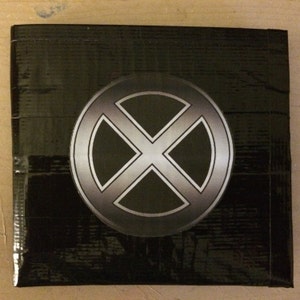 Puede incluir: Un objeto cuadrado negro con un logotipo de X-Men plateado y negro en el centro. El objeto parece estar hecho de un material texturizado, posiblemente cinta adhesiva o un adhesivo similar.