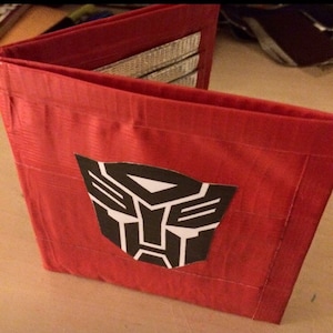Może przedstawiać: Czerwony portfel z czarno-białym logo Transformers. Portfel ma wiele przegródek i srebrne wnętrze. Portfel jest wykonany z teksturowanego materiału.