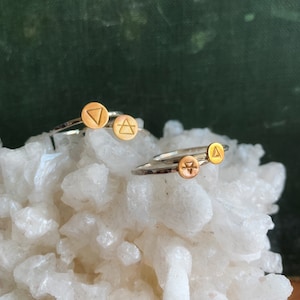May include: Three silver rings with gold accents. Each ring features a different geometric symbol. The rings are displayed on a white rock surface.