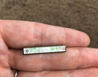 May include: A small, rectangular bar pin with a white enamel background and green floral design. The pin has a silver-toned metal base.