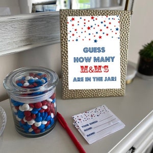 May include: A glass jar filled with red, white, and blue M&M's. A sign reads "Guess how many M&M's are in the jar!" with red, white, and blue stars in the background. There are also blank cards with the text "Your Guess", "Name", and "Name".