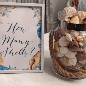 May include: A white framed sign with blue and gold watercolor seashells and seahorses reads "How Many Shells?"  A glass jar filled with seashells sits on a white surface.