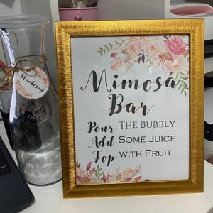 Op de afbeelding: Een gouden lijst met een bloemenontwerp en de tekst "Mimosa Bar" met instructies om "De bubbels te schenken, Sap toe te voegen, Top met fruit".