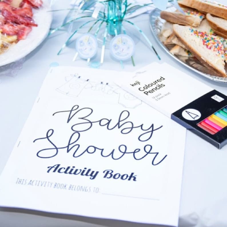 May include: A white activity book with the text "Baby Shower Activity Book" and the words "This activity book belongs to..." printed on the front cover. The book is lying on a white table with a box of coloured pencils and a plate of food in the background.