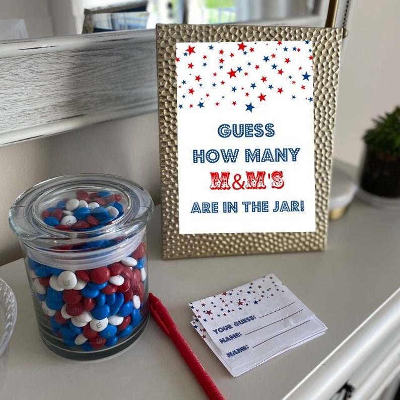 May include: A glass jar filled with red, white, and blue M&M's. A sign reads "Guess how many M&M's are in the jar!" with red, white, and blue stars in the background. There are also blank cards with the text "Your Guess", "Name", and "Name".