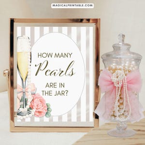 Op de afbeelding: Een ingelijst bord met de tekst "HOW MANY Pearls ARE IN THE JAR?" naast een glazen pot gevuld met parels. Het bord toont een champagneglas, roze strik en een roos. De pot is versierd met een roze strik.