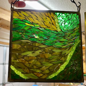 May include: A stained glass window panel with a mosaic design of green, yellow, and brown glass pieces. The glass pieces are arranged in a wave-like pattern.