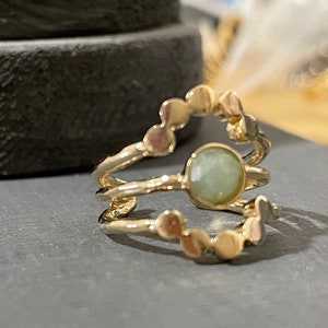 May include: A gold-colored ring with a light green gemstone at the center. The ring has a unique design with a series of small, circular accents. The ring is set against a dark gray surface.
