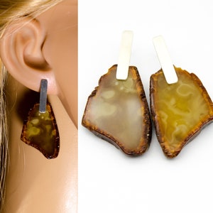 Puede incluir: Un par de pendientes de ámbar con postes de plata. El ámbar es de color marrón claro con un patrón de mármol. Los pendientes son una pieza de joyería única y elegante.