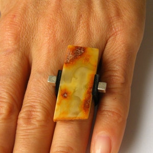 May include: A rectangular amber ring with black and silver accents. The ring is set on a finger.