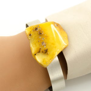 Könnte beinhalten: Ein Armreif mit einem großen, unregelmäßig geformten Bernstein. Der Bernstein hat eine warme gelbe Farbe mit dunkleren Einschlüssen. Das Armband besteht aus gebürstetem Metall. Ein einzigartiges Schmuckstück.
