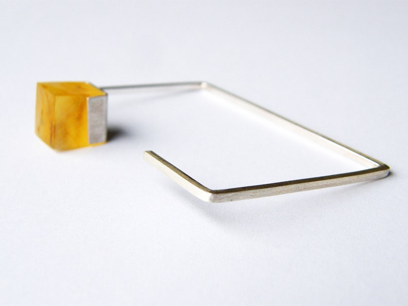 May include: A silver square hoop earring with a yellow amber cube attached to the top.