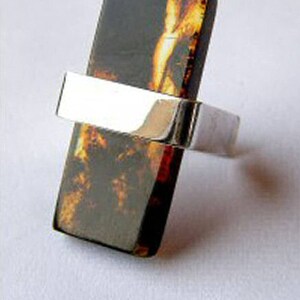 May include: A silver ring featuring a rectangular amber stone. The amber is a dark brown colour with streaks of orange and yellow.