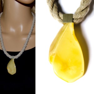 May include: A yellow amber pendant hangs from a thick, natural rope necklace. The pendant is teardrop shaped and has a smooth, polished surface.
