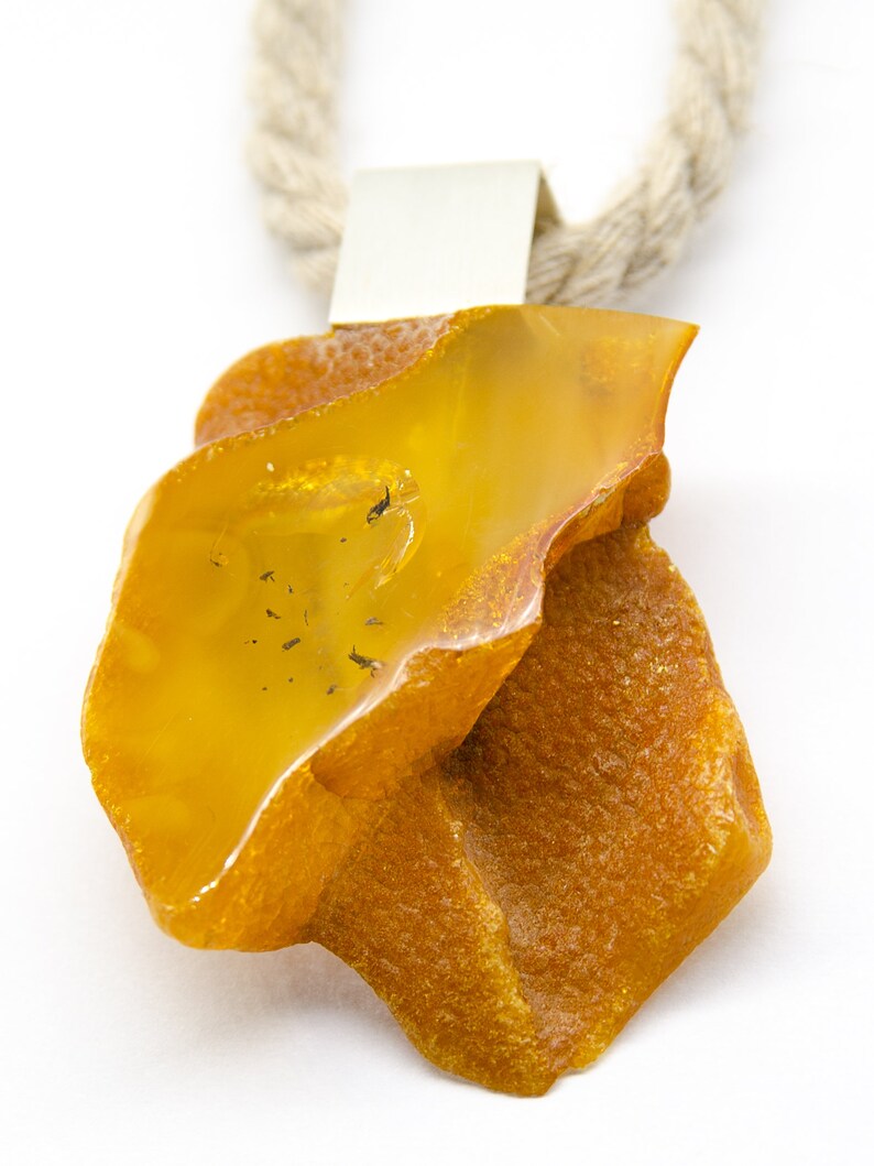Pode incluir: Um grande pingente de &acirc;mbar de forma irregular com uma argola de prata, pendurado em uma corda de fibra natural.