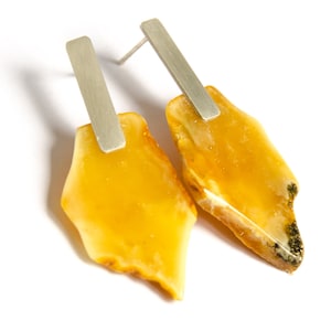 Puede incluir: Un par de pendientes de botón con piedras de ámbar amarillo translúcido de forma irregular, unidas a barras verticales de metal plateado cepillado. Las piedras tienen una superficie pulida con inclusiones naturales visibles.