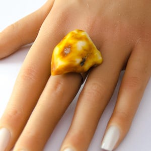May include: A ring featuring a large, irregularly shaped amber stone. The amber is a mix of yellow and white hues, with a polished surface. The ring's band is a simple silver color, and the amber stone is the focal point of the jewelry.
