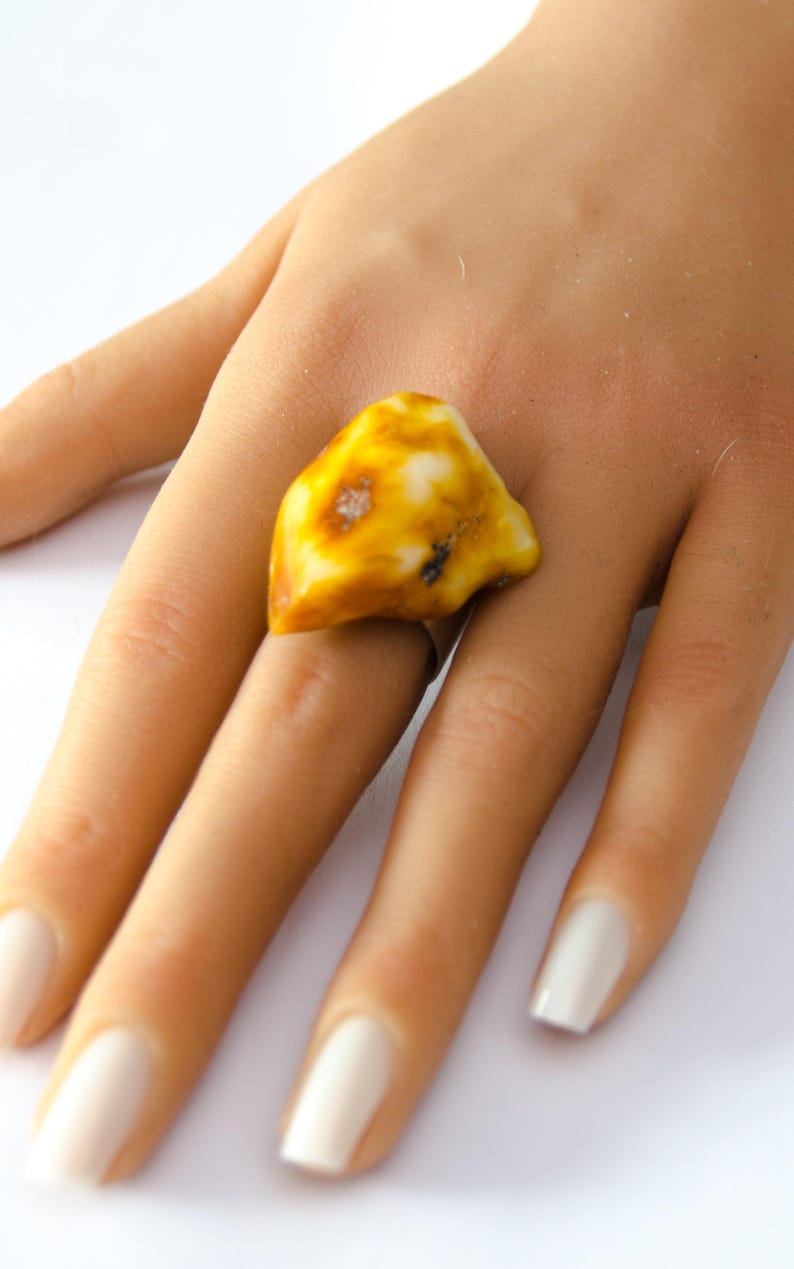 May include: A ring featuring a large, irregularly shaped amber stone. The amber is a mix of yellow and white hues, with a polished surface. The ring's band is a simple silver color, and the amber stone is the focal point of the jewelry.