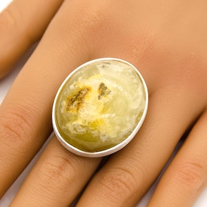 Könnte beinhalten: Ein ovaler Ring mit einem hellgrünen Edelstein mit braunen Einschlüssen, gefasst in einem silberfarbenen Metallband. Der Ring wird an einer Hand getragen. Der Edelstein ist durchscheinend. Modeschmuck.