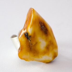 May include: A unique ring featuring a large, irregularly shaped amber stone. The amber is a warm, translucent yellow-orange color with white and dark brown inclusions. The ring's band is a simple, polished silver.