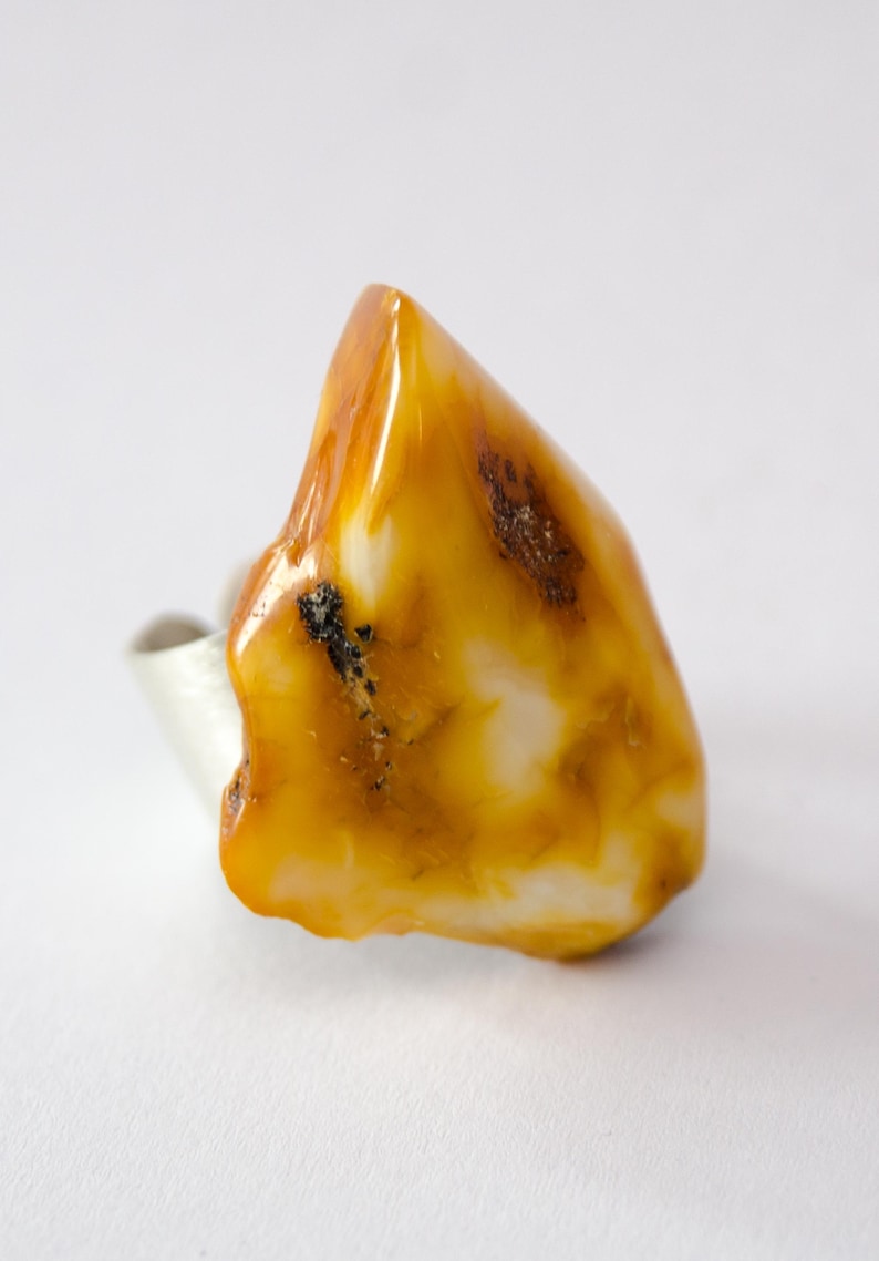 May include: A unique ring featuring a large, irregularly shaped amber stone. The amber is a warm, translucent yellow-orange color with white and dark brown inclusions. The ring's band is a simple, polished silver.