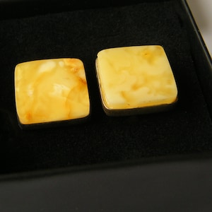 Peut inclure: Une paire de boutons de manchette carrés dotés de pierres d'ambre polies, translucides, de couleur jaune et crème, serties dans un cadre métallique simple. Les boutons de manchette sont présentés côte à côte dans une boîte noire.