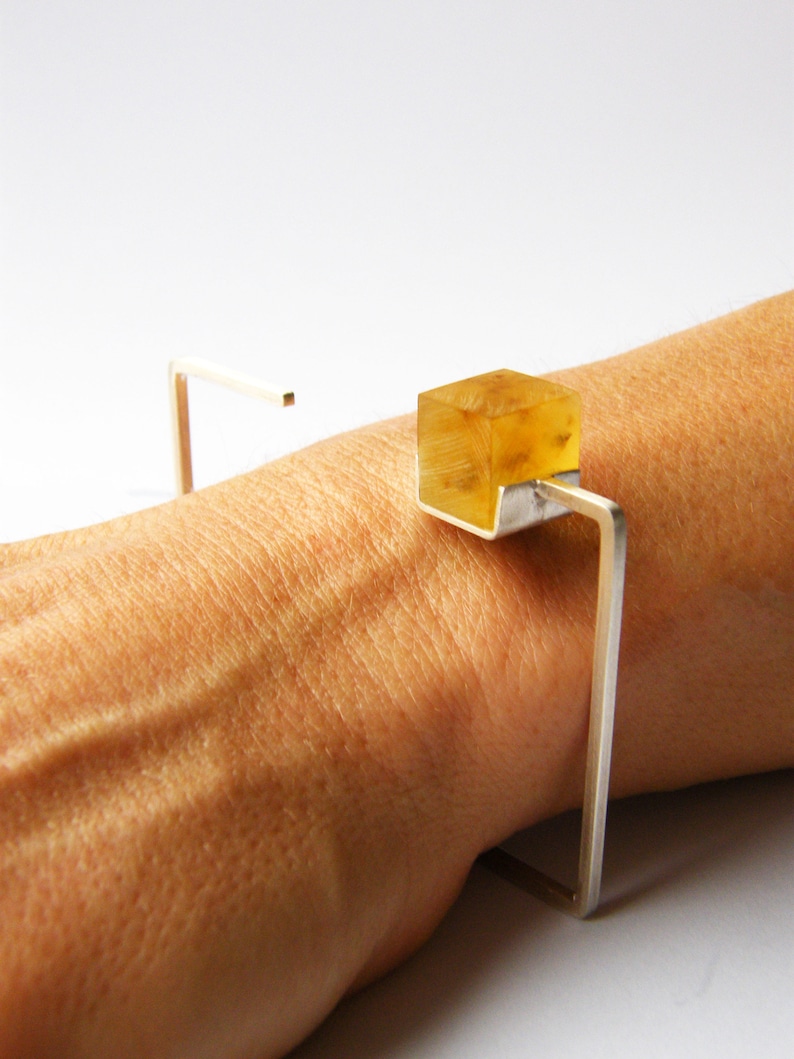 May include: A silver bracelet with a clear, yellow, cube-shaped stone. The bracelet is open and has a simple, geometric design.