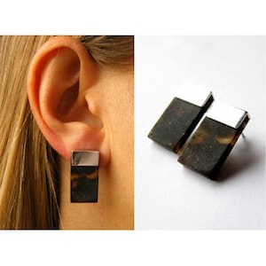 Puede incluir: Un par de pendientes rectangulares de plata y marrón. Los pendientes tienen una parte superior plateada y una parte inferior marrón. La parte inferior marrón está hecha de un material natural, como madera o piedra.