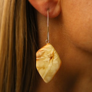 Puede incluir: Un par de pendientes colgantes con una piedra de ámbar marrón claro en forma de diamante. Los pendientes son de plata y tienen un diseño sencillo.