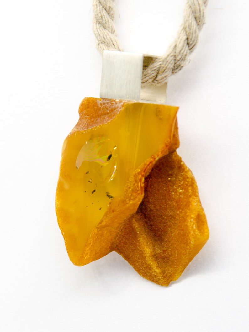 Pode incluir: Um pingente de &acirc;mbar amarelo com uma argola de prata, pendurado em uma corda de fibra natural.
