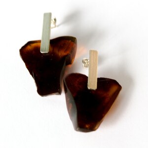 Puede incluir: Un par de pendientes de color ámbar con formas irregulares y barras plateadas. Los pendientes tienen un acabado pulido y brillante y están sobre un fondo blanco. Una pieza de joyería única.