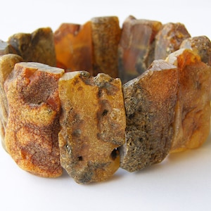 Puede incluir: Una pulsera elástica compuesta por múltiples piezas rectangulares de ámbar en bruto y sin pulir. Cada segmento presenta una textura rugosa única con tonos variados de dorado, naranja y marrón oscuro, mostrando la forma orgánica natural de la resina.