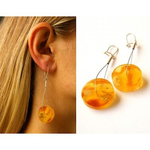 Puede incluir: Un par de pendientes de ámbar con alambre de plata y cierres de gancho. El ámbar es de un color miel cálido y tiene un aspecto natural y orgánico.