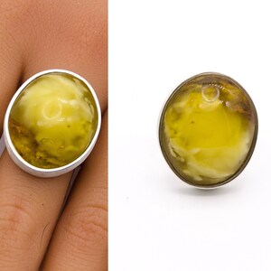 May include: A silver ring featuring a large, oval-shaped amber stone. The amber has a translucent, yellowish-green hue with internal patterns. The ring is shown on a finger and also isolated against a white background.