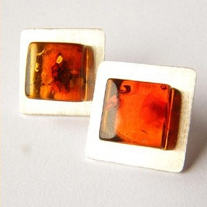 Puede incluir: Un par de pendientes de plata con piedras de ámbar cuadradas. El ámbar es de color naranja intenso con matices marrones y rojos.