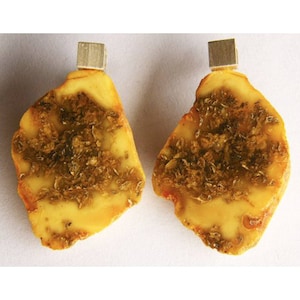 Puede incluir: Un par de pendientes de ámbar con un cierre de plata. El ámbar es de color amarillo claro con inclusiones marrones.