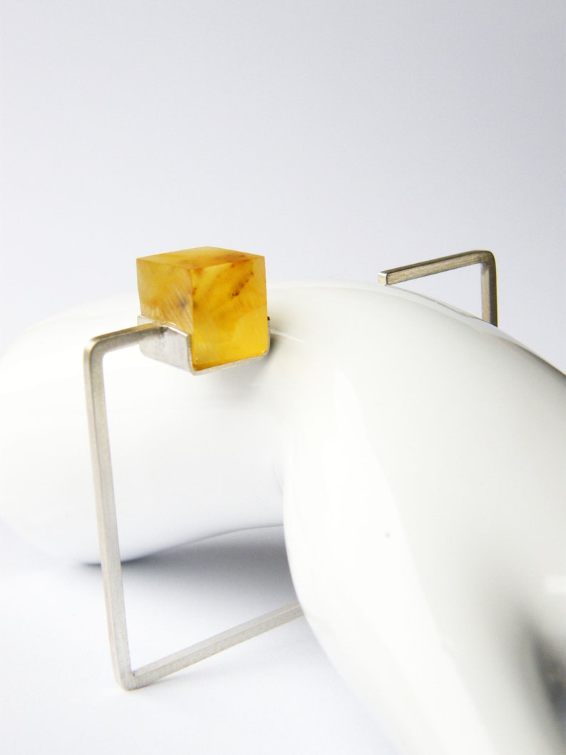 May include: A silver ring with a square amber stone set in the center. The ring is on a white surface.