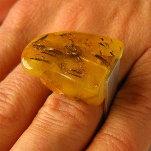 Puede incluir: Un anillo con una piedra grande de ámbar amarillo con inclusiones negras. La piedra está engastada en una banda de plata.