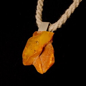 Pode incluir: Um colar com um pingente de &acirc;mbar de forma irregular, engastado em prata. O colar &eacute; feito com um cord&atilde;o grosso de corda natural.